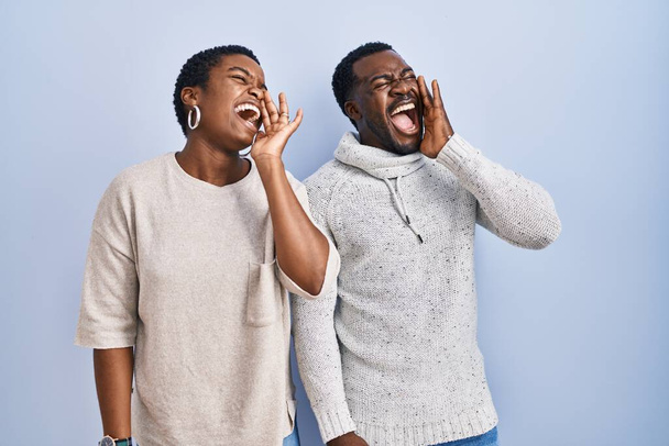 Genç Afro-Amerikan çifti mavi arka planda yan yana durup el ele bağırıyor ve çığlık atıyorlar. iletişim konsepti.  - Fotoğraf, Görsel