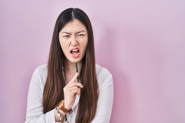 Chinese young woman holding pencil over pink background angry and mad screaming frustrated and furious, shouting with anger. rage and aggressive concept.  - Fotografie, Obrázek