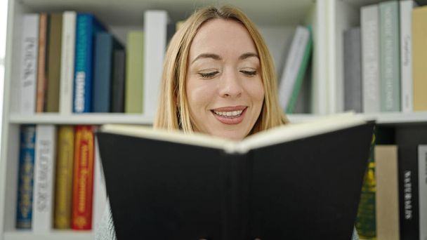 Junge blonde Studentin lächelt selbstbewusst beim Lesen von Büchern in der Bibliothek - Foto, Bild