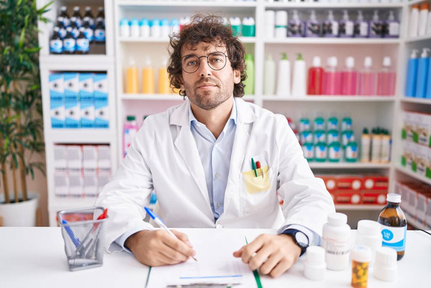 Ισπανόφωνος νεαρός άνδρας που εργάζεται στο φαρμακείο χαλαρός με σοβαρή έκφραση στο πρόσωπο. απλή και φυσική κοιτάζοντας την κάμερα.  - Φωτογραφία, εικόνα
