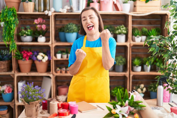 Латиноамериканка средних лет, работающая в цветочном магазине, радуется удивлению и удивлению за успех с поднятыми руками и закрытыми глазами  - Фото, изображение