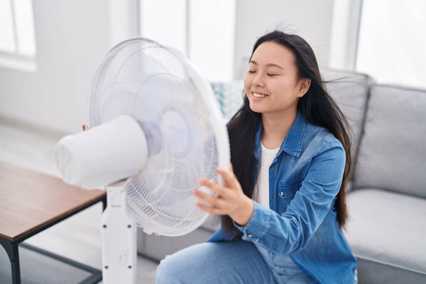 Молодая китаянка с помощью вентилятора сидит дома на диване - Фото, изображение