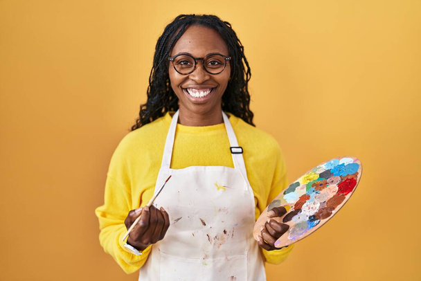 Αφρικανή που κρατάει την παλέτα του ζωγράφου με ένα χαρούμενο και δροσερό χαμόγελο στο πρόσωπο. τυχερό άτομο.  - Φωτογραφία, εικόνα