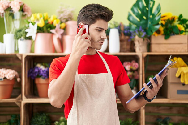 Молодий іспаномовний чоловік флорист розмовляє на смартфоні читаючи документ у квітковому магазині
 - Фото, зображення