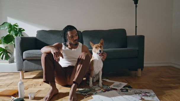 Θετικός άντρας που παριστάνει το κατοικίδιο στο σπίτι. Αφρο-αμερικάνος που αγκαλιάζει το σκύλο που κάθεται στο πάτωμα. Χαμογελώντας καλλιτέχνης χαλάρωση μετά από σχέδιο closeup. Σύγχρονη ζωγράφος αναπαύεται με κατοικίδια ζώα - Φωτογραφία, εικόνα