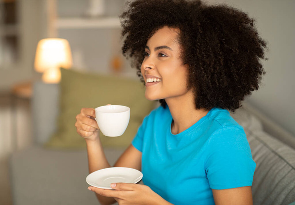Rire joyeuse jeune femme bouclée noire boit une tasse de café chaud sur le canapé, profite de temps libre seul le soir, se détendre ou bonjour dans le salon intérieur. Temps de thé et repos à la maison - Photo, image