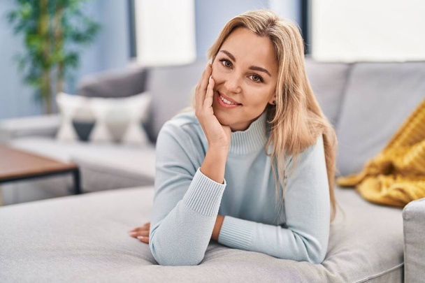 Nuori blondi nainen hymyilee luottavainen makaa sohvalla kotona - Valokuva, kuva