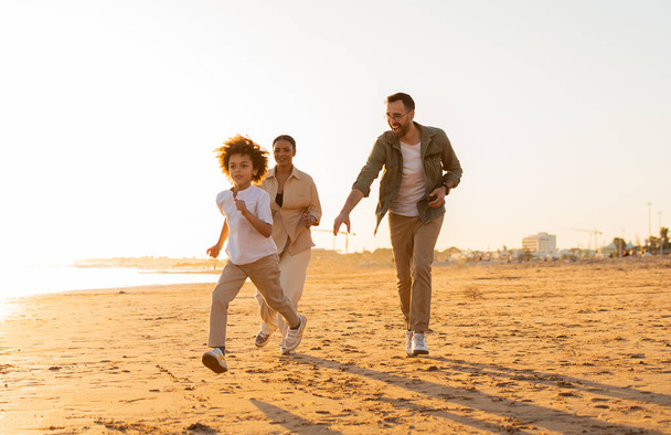 Zabawa na plaży. Szczęśliwa, różnorodna rodzina spacerująca i wygłupiająca się nad morzem, chłopiec biegający wzdłuż plaży ścigany przez rodziców, pełna długość, wolna przestrzeń - Zdjęcie, obraz