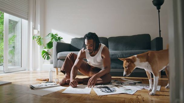 Inspired painter creating artwork in workshop. Domestic dog looking at pictures. Dreadlocks young man drawing black chalk in studio. Talented african american artist working masterpiece in workplace  - Foto, Imagem
