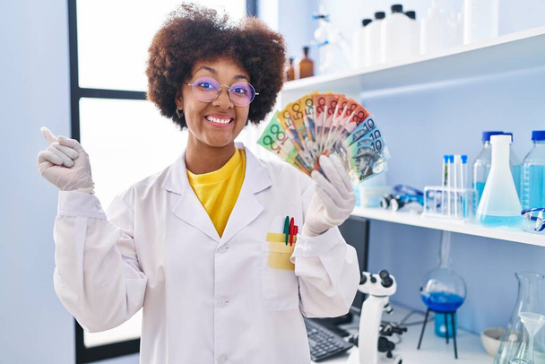 Молодая африканская американка, работающая в научной лаборатории, держит деньги, улыбаясь, показывая пальцем и рукой в сторону.  - Фото, изображение