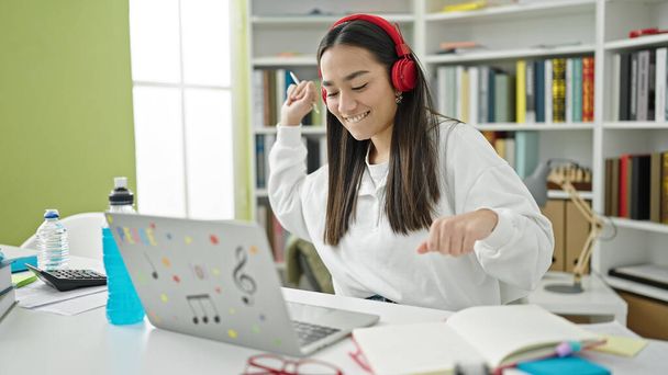 Jovem bela estudante hispânica usando laptop e fones de ouvido dançando na sala de aula da universidade - Foto, Imagem