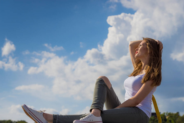 Trendy fashionable woman relaxing outdoor wearing casual footwear white sneakers and hole trousers. Female enjoy sunlight against blue sky. - Fotografie, Obrázek