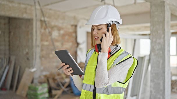 Joven mujer rubia arquitecta hablando en el teléfono inteligente mirando touchpad en el sitio de construcción - Foto, Imagen