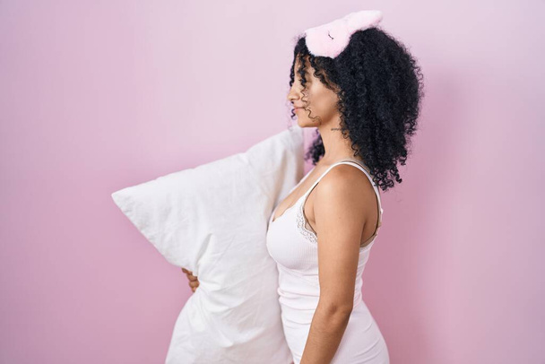 Latynoska kobieta z kręconymi włosami nosząca maskę snu i piżamę trzymająca poduszkę patrzącą na bok, relaksująca się poza profilem z naturalną twarzą i pewnym uśmiechem.  - Zdjęcie, obraz