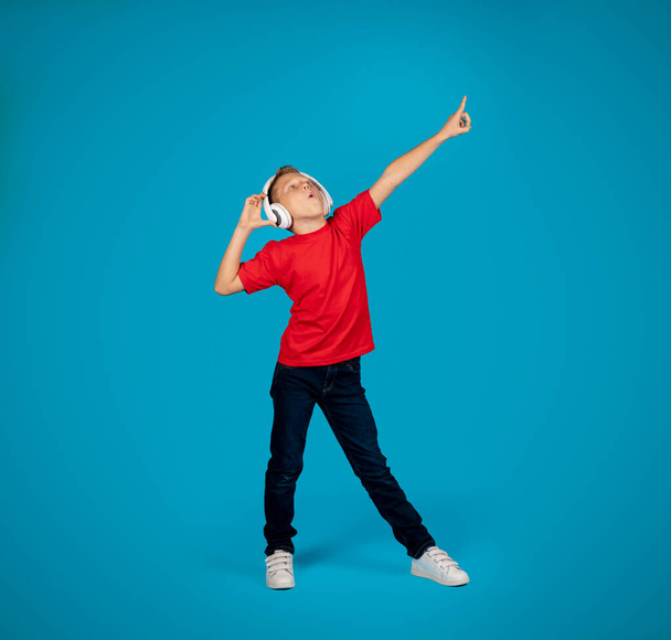 Cheerful Caucasian Boy Wearing Wireless Headphones Listening Music And Dancing, Preteen Male Child Having Fun While Standing Over Blue Background In Studio, Full Length Shot, Copy Space - Foto, Imagen