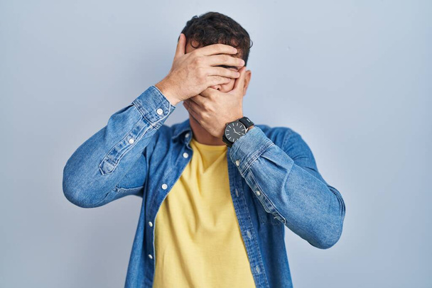 Young hispanic man standing over blue background covering eyes and mouth with hands, surprised and shocked. hiding emotion  - 写真・画像