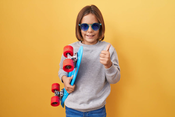 Pequeño niño hispano con gafas de sol sosteniendo patinaje sonriendo feliz y positivo, pulgar hacia arriba haciendo excelente y signo de aprobación  - Foto, Imagen