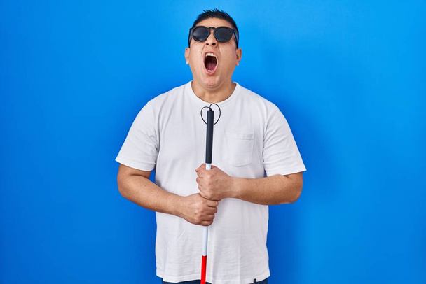 Hispanic young blind man holding cane angry and mad screaming frustrated and furious, shouting with anger looking up.  - Foto, afbeelding