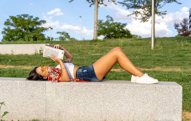 Χαλαρή γυναίκα με γυαλιά ηλίου που διαβάζει ένα βιβλίο ενώ ξαπλώνει σε ένα παγκάκι σε ένα πάρκο. - Φωτογραφία, εικόνα