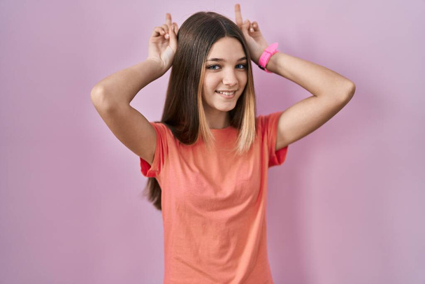 Teenager girl standing over pink background posing funny and crazy with fingers on head as bunny ears, smiling cheerful  - Valokuva, kuva