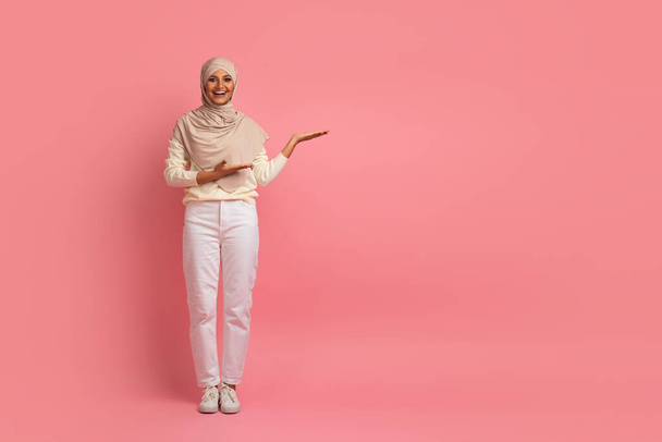 Nice Promo. Smiling Muslim Woman In Hijab Poiting Aside At Copy Space With Two Hands, Cheerful Young Arabic Woman In Headscarf Indicating Free Space For Advertisement, Standing On Pink Background - Foto, Bild