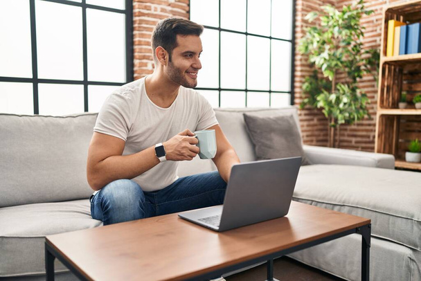 Young hispanic man using laptop and drinking coffee sitting on sofa at home - Foto, Imagen