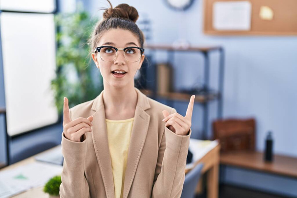 Adolescente travaillant au bureau portant des lunettes étonnée et surprise regardant vers le haut et pointant avec les doigts et les bras levés.  - Photo, image