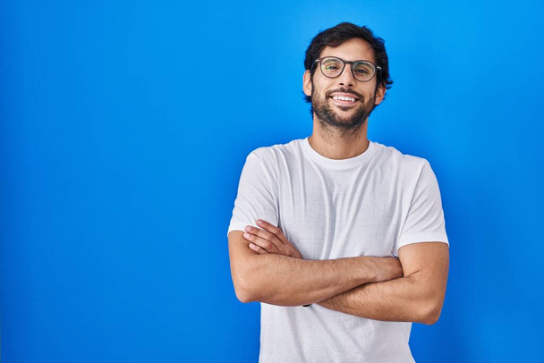 Handsome latin man standing over blue background happy face smiling with crossed arms looking at the camera. positive person.  - Fotoğraf, Görsel