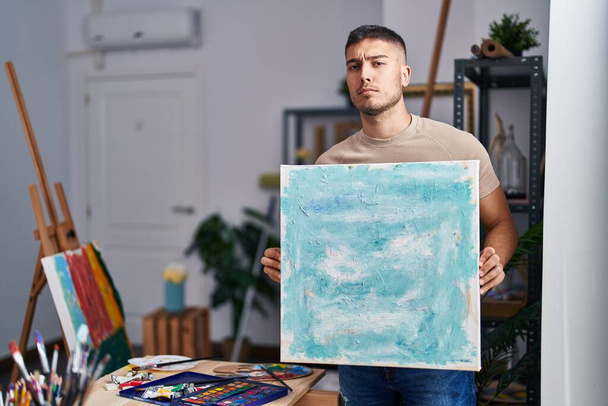 Joven pintor hispano hombre sosteniendo lienzo escéptico y nervioso, frunciendo el ceño molesto debido a un problema. persona negativa.  - Foto, Imagen