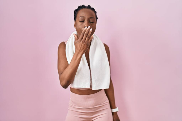 Piękna czarna kobieta ubrana w odzież sportową i ręcznik na różowym tle znudzona ziewaniem zmęczona przykrywaniem ust dłonią. niepokój i senność.  - Zdjęcie, obraz