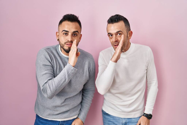 Homosexuelles Paar steht über rosa Hintergrund Hand auf Mund und erzählt geheime Gerüchte, flüstert bösartige Gespräche  - Foto, Bild