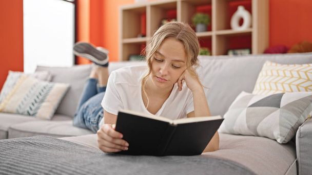 Young blonde woman reading book lying on sofa at home - Foto, Imagem