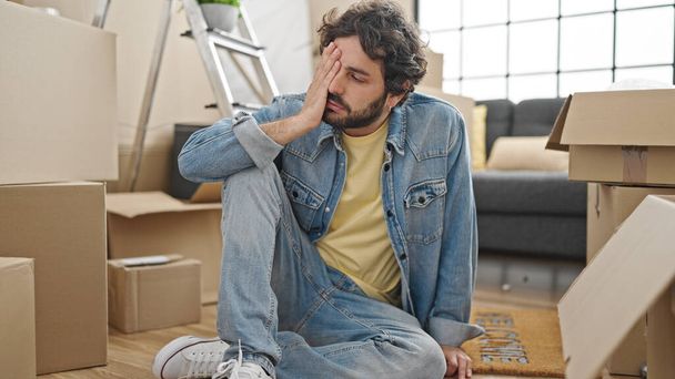 Young hispanic man sitting on floor looking upset at new home - 写真・画像