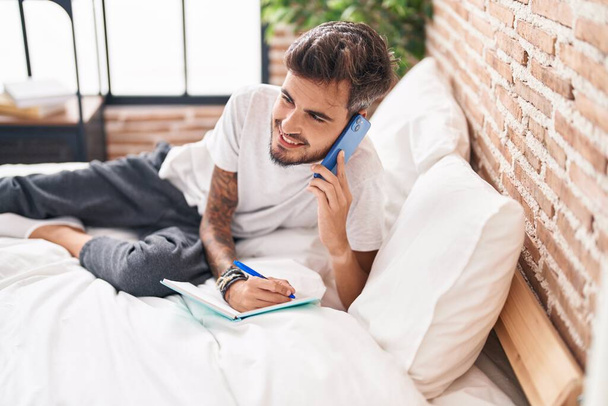 Joven hispano hablando en un smartphone escribiendo en un libro en el dormitorio - Foto, Imagen