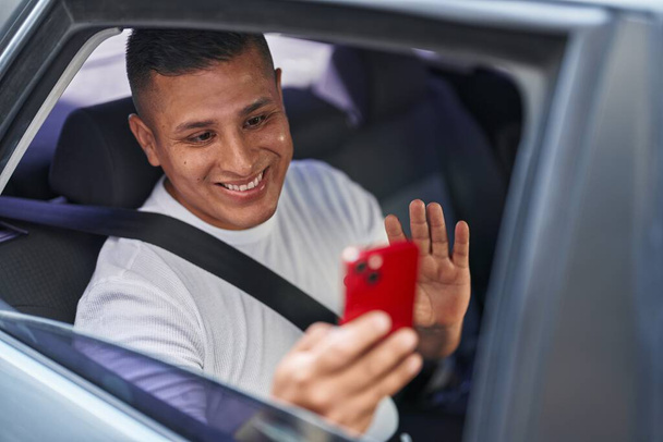 Jeune homme hispanique faire appel vidéo avec smartphone dans la voiture regardant positif et heureux debout et souriant avec un sourire confiant montrant les dents  - Photo, image