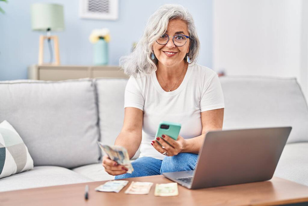 Middle age woman accounting using laptop and smartphone sitting on sofa at home - Photo, Image