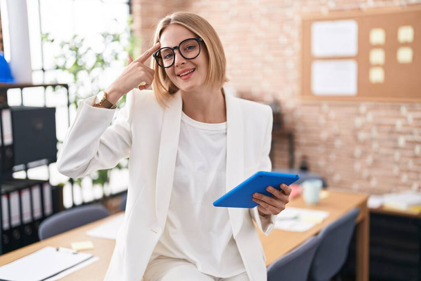 Молодая белая женщина работает в офисе в очках, улыбаясь, указывая на голову одним пальцем, отличная идея или мысль, хорошая память  - Фото, изображение