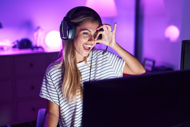 Молодая блондинка, играющая в видеоигры улыбаясь счастливая делает хорошо знак с рукой на глазу, глядя сквозь пальцы  - Фото, изображение