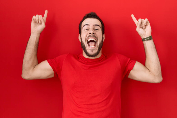 Nuori latinomies yllään rento punainen t paita hymyillen hämmästynyt ja yllättynyt ja osoittaa ylös sormilla ja nosti kädet.  - Valokuva, kuva