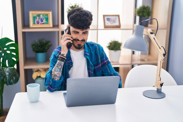 Hombre hispano joven usando portátil y hablando en el teléfono inteligente en casa - Foto, imagen