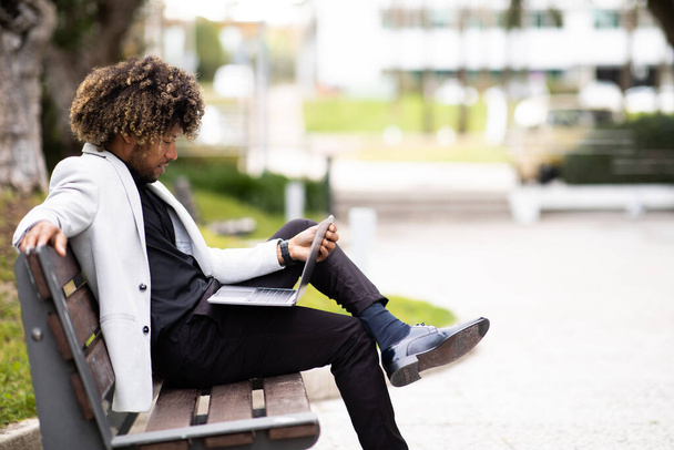 Black businessman in suit using laptop while sitting on bench near modern office building, side view, free space. Conference due social distance, new normal concept - Фото, изображение