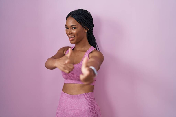 Mujer afroamericana con trenzas que usan ropa deportiva sobre fondo rosa señalando los dedos a la cámara con la cara feliz y divertida. buena energía y vibraciones.  - Foto, imagen