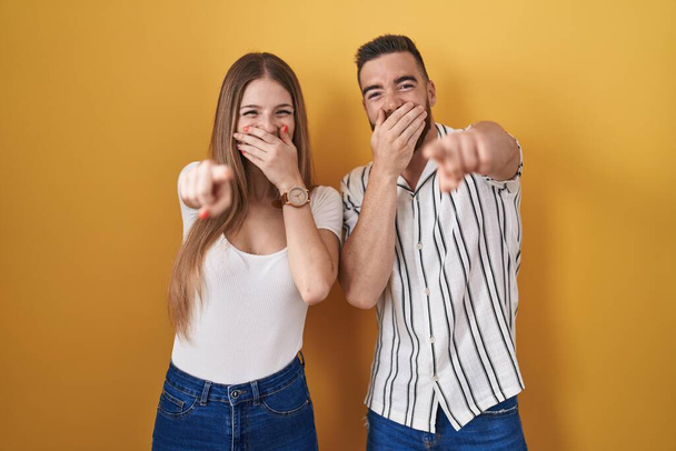 Young couple standing over yellow background laughing at you, pointing finger to the camera with hand over mouth, shame expression  - Photo, Image