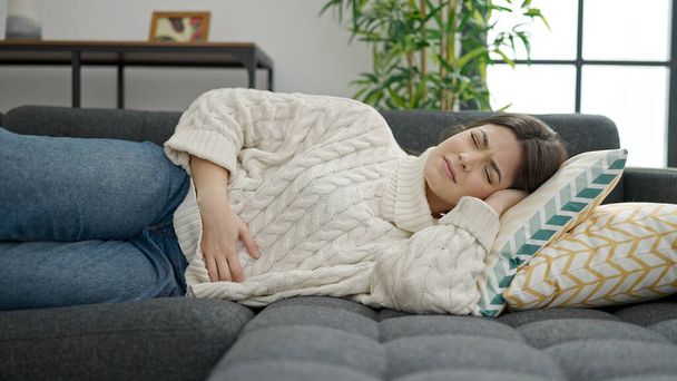 Joven mujer hispana hermosa que sufre de dolor de estómago tumbada en el sofá en casa - Foto, Imagen