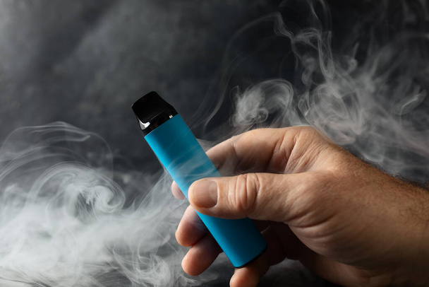 eldobható elektronikus cigaretta egy férfi kezében egy sötét háttérben, füsttel körülvéve. A modern alternatív dohányzás, a gőzölés és a nikotin fogalma - Fotó, kép