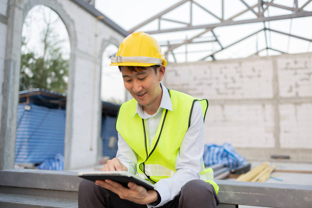Happy Asian smiling in engineering uniform at construction site while holding a tablet. Young professional foreman working on structure building at workplace - with copy space - 写真・画像