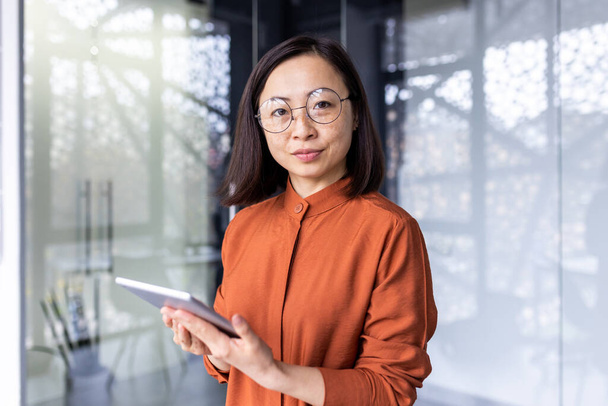 Portrait of a young Asian woman programmer, developer, tester in glasses standing in the office, holding a tablet, working online and looking, posing at the camera. - Фото, зображення