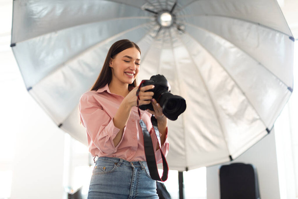 Jovem fotógrafa trabalhando com câmera profissional na frente do guarda-chuva reflexivo, tirando fotos e desfrutando de seu trabalho em photostudio moderno, espaço de cópia - Foto, Imagem