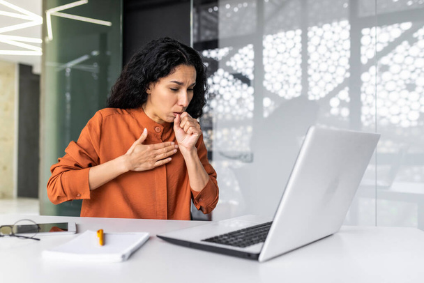 Attack of cough, asthma, allergies at work. Young hispanic woman sitting in office at work, covering mouth with hand, holding chest, suffering from cough, heavy breathing. - Photo, image