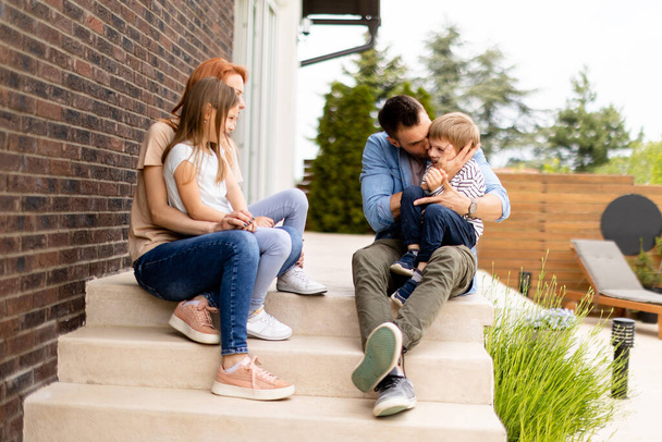 Family with a mother, father son and daughter sitting outside on  steps of a front porch of a brick house - Photo, Image